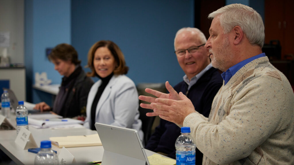 Jeff Frost and Andrea Ball sit at a table smiling and looking to their left at a caucasian man with grayish-white hair and a beard. He is talking and gesturing with his hands.