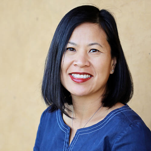 Close-up of Jackie Thu-Huong, an Asian woman with shoulder-length black hair, smiling at the camera. She wears dark red lipstick and a blue top.