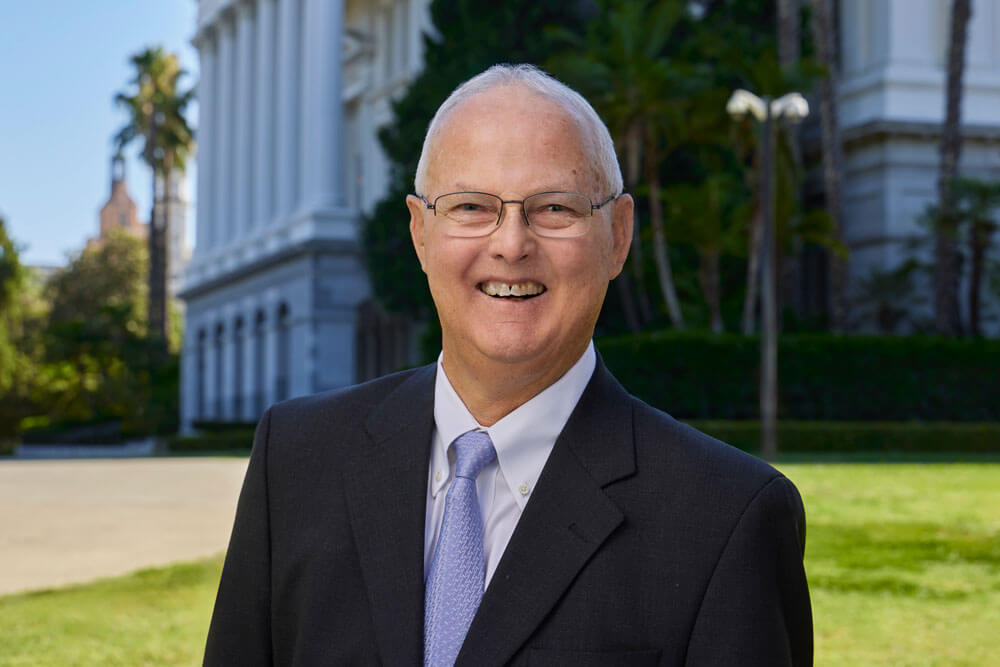 Jeff Frost, a caucasian man with white hair and glasses, smiles and wears a dark suit, white collared shirt, and lavender tie. The Sacramento Capitol building is in the background.