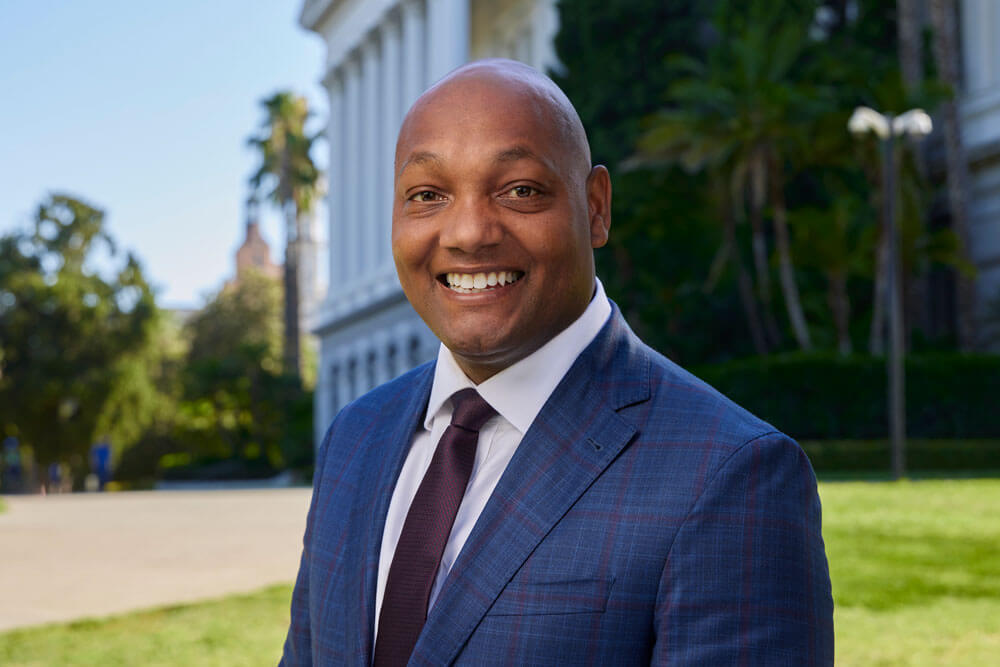 Khieem Jackson, a bald black man, smiles and wears a dark purple and blue patterned suit, white collared shirt, and dark purple tie. The Sacramento Capitol building is in the background.