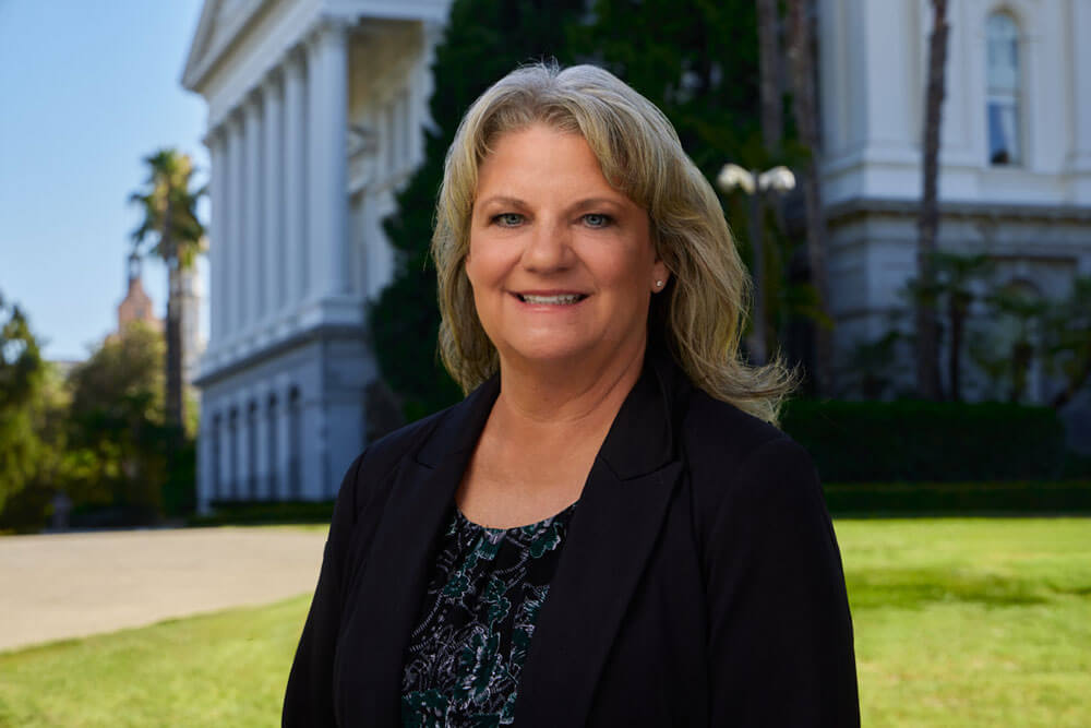 Laura Wasco, a caucasian woman with medium-length dark blonde hair, smiles and wears a dark suit jacket with a dark multi-colored top. The Sacramento Capitol building is in the background.