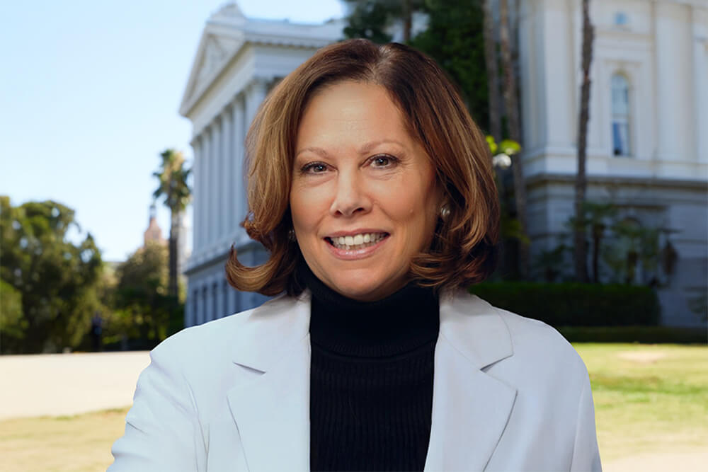 Andrea Ball, a caucasian woman with medium-length brown hair, smiles and wears a light suit jacket with a dark turtleneck top. The Sacramento Capitol building is in the background.