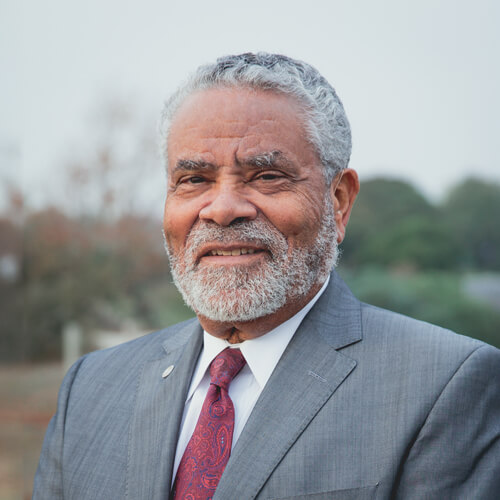 Carl Cohn, an older medium to dark-skinned man with gray hair and beard, smiles at the camera. He wears a gray suit, white dress shirt, and a red and blue paisley-patterned tie.