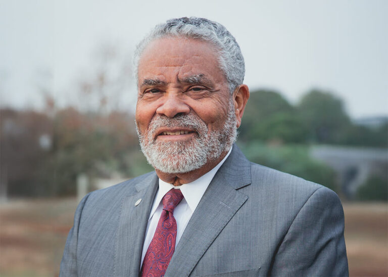 Carl Cohn, an older medium to dark-skinned man with gray hair and beard, smiles at the camera. He wears a gray suit, white dress shirt, and a red and blue paisley-patterned tie.