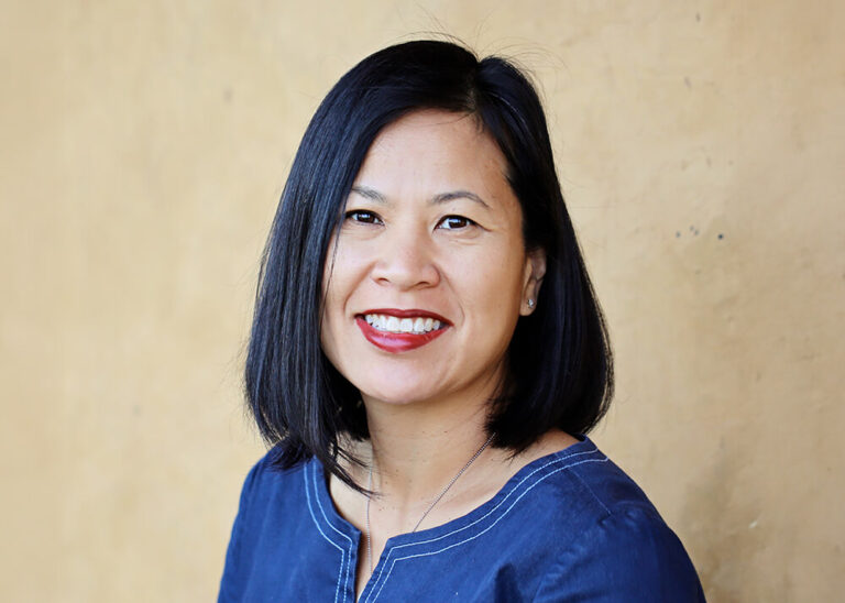 Close-up of Jackie Thu-Huong, an Asian woman with shoulder-length black hair, smiling at the camera. She wears dark red lipstick and a blue top.